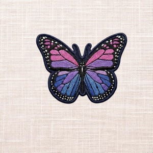 May include: Embroidered butterfly patch with a navy blue outline. The butterfly's wings feature a gradient of colors, including pink, purple, and blue. White dots line the edges of the wings. The patch is likely for clothing or accessories.