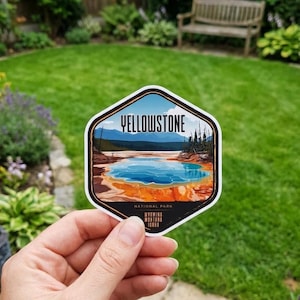 May include: A hexagonal sticker featuring a Yellowstone National Park landscape. The sticker displays a blue hot spring, mountains, and trees under a blue sky. The word "YELLOWSTONE" is at the top, with "NATIONAL PARK" and the states listed at the bottom.