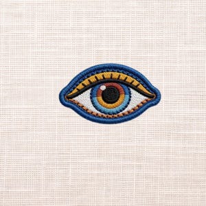 May include: Embroidered eye patch featuring a detailed design with a blue, yellow, orange, and white color scheme. The eye has a black pupil and is outlined in blue. The patch is likely for clothing or accessories.