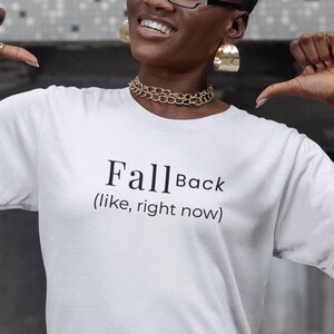 Könnte beinhalten: Weißes T-Shirt mit dem schwarzen Text "Fall Back (like, right now)". Das Model trägt eine Brille, Goldschmuck und eine blaue Jeans.