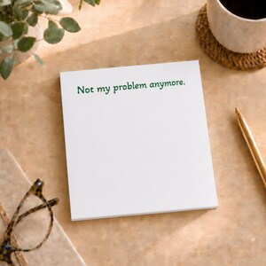 May include: A white rectangular notepad featuring the dark green printed text Not my problem anymore. The notepad is positioned on a light brown surface alongside a pair of tortoiseshell glasses, a gold pen, and a white ceramic mug on a coaster.