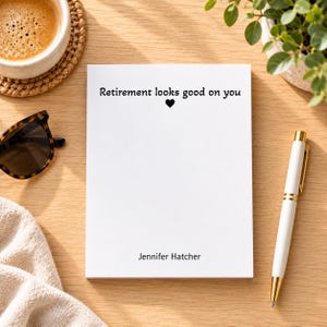 May include: A white rectangular notepad featuring the black printed text Retirement looks good on you with a small heart icon below it and the name Jennifer Hatcher printed in black at the bottom of the page.