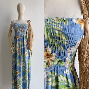 Puede incluir: Vestido largo azul claro con estampado floral. El vestido presenta un corpiño fruncido y tirantes finos. El estampado floral incluye flores y hojas blancas, amarillas y verdes. Un bolso de macramé beige está colgado sobre el hombro del maniquí.
