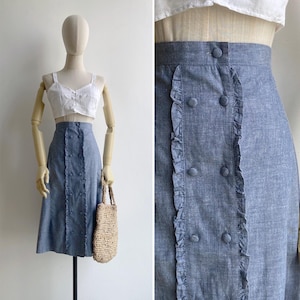 May include: A denim skirt with a ruffled trim and button detailing. The skirt is a mid-length style and is light blue in colour.