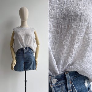 May include: A white textured sleeveless top, knotted at the waist, and blue denim shorts. The shorts have a button closure and distressed details. The mannequin is on a black stand against a white background.