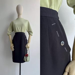 May include: A black pencil skirt with button details on the side, paired with a light green long-sleeved top. The skirt has a tailored fit and a classic design. A small, light green purse with a red flower is also visible.