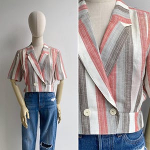 Op de afbeelding: Een gestreepte blazer met korte mouwen in rood, wit en grijs. De blazer heeft een dubbele rij knopen met twee witte knopen en een revers. Het kledingstuk wordt op een mannequin getoond.