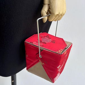 Puede incluir: Un bolso de mano rojo y geométrico con asa plateada. El bolso presenta un estampado floral y una base beige. El bolso es sostenido por una mano de madera.