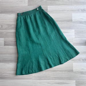 Könnte beinhalten: Ein langer, smaragdgrüner Rock mit hoher Taille und Knopfverschluss. Der Rock ist aus einem strukturierten Stoff gefertigt und hat einen leichten Schlag am Saum. Der Rock ist einfarbig und scheint aus Leinen zu sein.