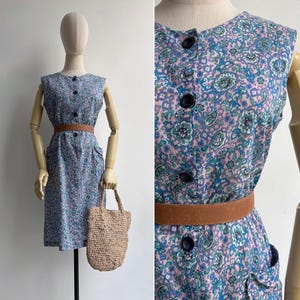 Pode incluir: Um vestido sem mangas com botões e um padrão floral em tons de azul, verde e roxo. O vestido apresenta um cinto castanho e dois bolsos frontais. Uma sacola de mão tecida é segurada pelo manequim. O vestido é exibido em um manequim.