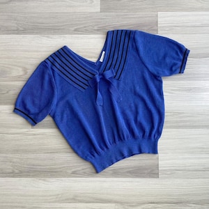 Peut inclure: Un haut en tricot à manches courtes bleu roi doté d'un large col style marin avec des rayures horizontales noires. Le vêtement comprend un ruban bleu assorti noué au centre de l'encolure et un liseré noir sur les poignets et l'ourlet.