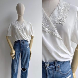 A mannequin wears a vintage 1980s white drapey crossover v-neck wrap top in white with a pretty & feminine embroidered lace collar, short sleeves & pleating at the bodice. It is shown tucked into a pair of high waisted blue jeans.