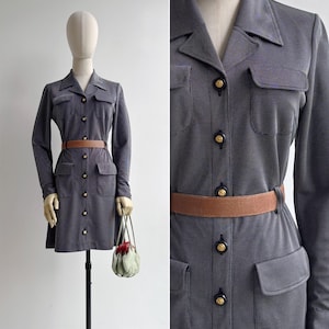 Könnte beinhalten: Ein dunkelgraues Langarm-Hemdkleid mit feinem Nadelstreifenmuster, spitzem Kragen und durchgehender Knopfleiste mit goldfarbenen runden Knöpfen. Das Kleid hat vier Pattentaschen und wird in der Taille mit einem hellbraunen Ledergürtel betont.