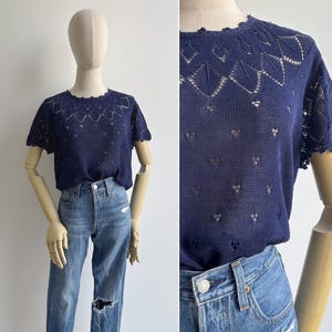 May include: A dark blue short-sleeved knit top featuring an intricate openwork diamond pattern across the chest and shoulders, paired with light blue distressed denim jeans that have a visible rip on the left knee and a metal button closure.