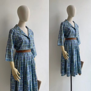 May include: A vintage two-piece dress set in blue, green, and white geometric print. The set includes a collared, button-down top with three-quarter sleeves and a pleated skirt. A brown belt cinches the waist. The dress is displayed on a mannequin.