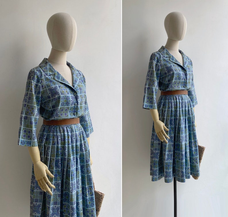 May include: A vintage two-piece dress set in blue, green, and white geometric print. The set includes a collared, button-down top with three-quarter sleeves and a pleated skirt. A brown belt cinches the waist. The dress is displayed on a mannequin.