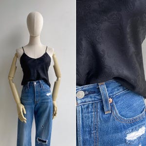 A mannequin wears a 90s silky cami tank with a woven paisley texture tucked not a pair of high waisted blue jeans
