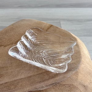 May include: A clear, decorative glass dish with a textured, organic design. The dish is shaped like a leaf or a natural form, with a rippled surface. The dish is resting on a wooden surface.