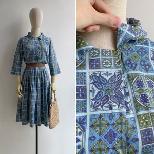 May include: A vintage blue dress with a matching top and pleated skirt, featuring a geometric floral pattern in shades of blue, green, and purple. The dress has a collared neckline, three-quarter sleeves, and a brown belt. A straw handbag completes the look.