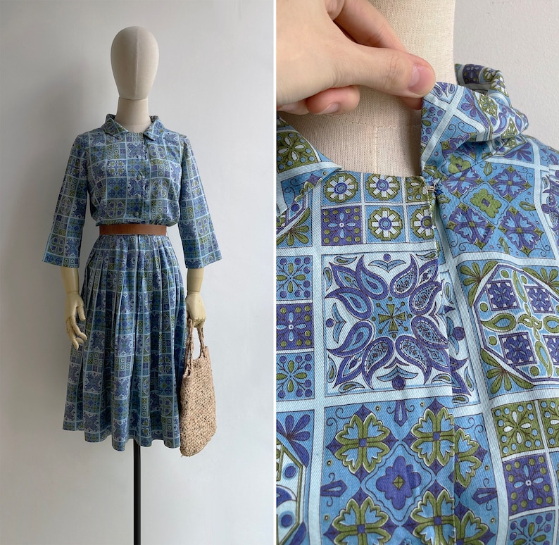 May include: A vintage blue dress with a matching top and pleated skirt, featuring a geometric floral pattern in shades of blue, green, and purple. The dress has a collared neckline, three-quarter sleeves, and a brown belt. A straw handbag completes the look.