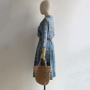 May include: A vintage blue dress with a square pattern, long sleeves, and a collared neckline. The dress is cinched at the waist with a brown belt. A woven straw tote bag is held by the mannequin. The dress is knee-length.