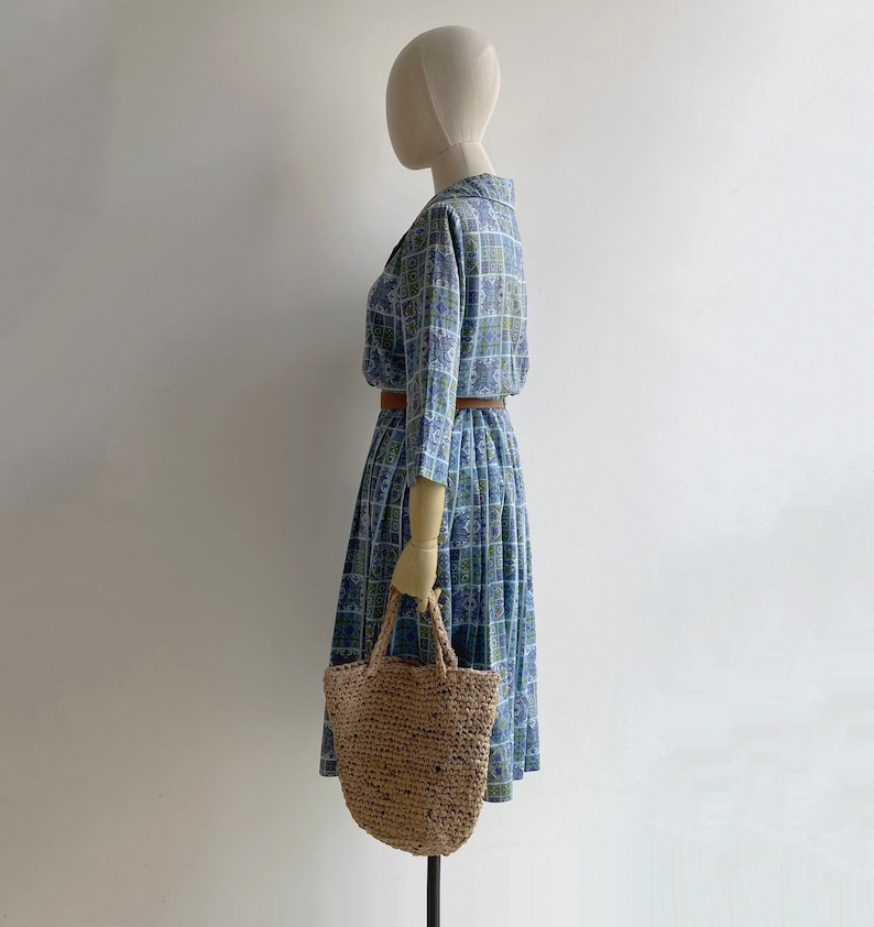 May include: A vintage blue dress with a square pattern, long sleeves, and a collared neckline. The dress is cinched at the waist with a brown belt. A woven straw tote bag is held by the mannequin. The dress is knee-length.