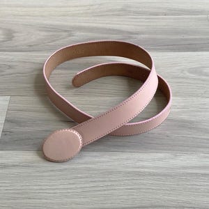 May include: A light pink belt featuring a smooth, matte finish and a circular buckle of the same colour. The belt strap has visible tonal stitching along the edges, and the interior side shows a tan-coloured lining with embossed text.