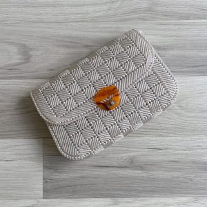 Puede incluir: Un bolso de mano de macramé gris claro con un patrón geométrico. El bolso tiene un cierre de solapa y un cierre de color carey. La bolsa está hecha de material tejido y está colocada sobre una superficie de madera gris claro.