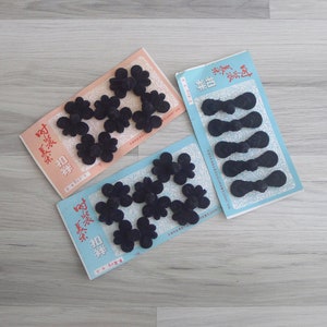 May include: Three packages of black fabric closures. Two packages feature flower-shaped closures, while the third has bow-tie style closures. The packages are on a light gray wooden surface. Text in Chinese characters is visible on the packaging.
