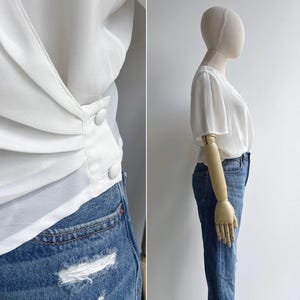 Side view of a mannequin in a retro 80s white polyester chiffon draped top with a feminine sheer lace collar. The left side of the image shows a close-up of the pleated waist detail & buttons.