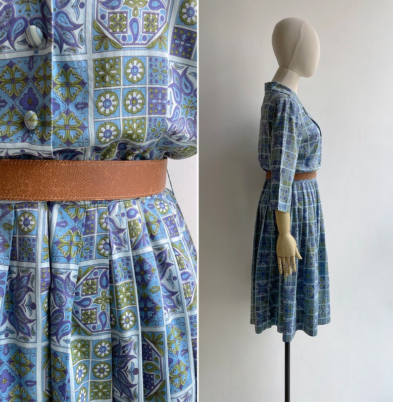 May include: A vintage dress with a blue, green, and purple geometric pattern. The dress features a button-down top, a pleated skirt, and a brown belt. The dress is displayed on a mannequin.