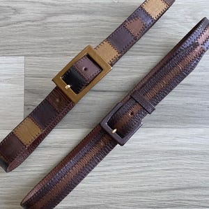 Op de afbeelding: Twee bruine leren riemen met rechthoekige gespen. Eén riem heeft een patchwork-ontwerp in bruin- en tinten. De andere riem heeft een gestreept patroon in vergelijkbare aardetinten. Beide riemen worden gepresenteerd op een lichtgrijze houten ondergrond.