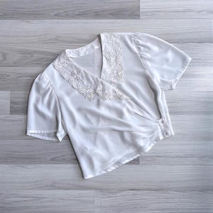 Flat lay of a vintage 80s white crossover wrap blouse with a floral embroidered lace collar, pleats across the waist & short sleeves.