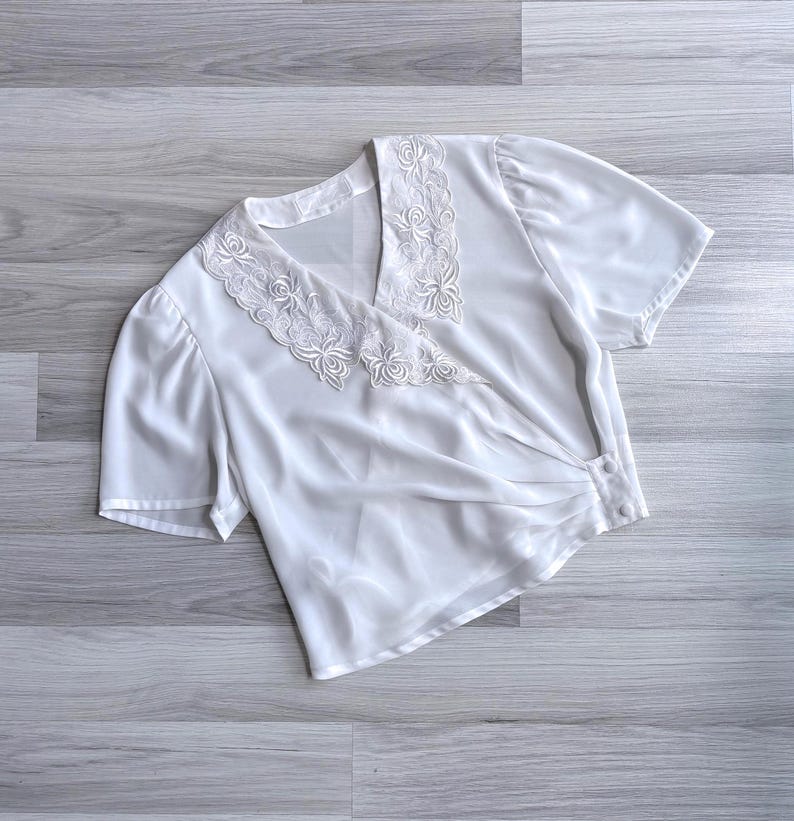 Flat lay of a vintage 80s white crossover wrap blouse with a floral embroidered lace collar, pleats across the waist & short sleeves.