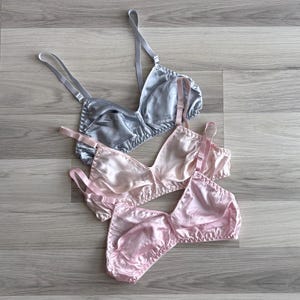 Könnte beinhalten: Drei BHs aus Satin in Silber- und Rosatönen sind auf einer Holzoberfläche angeordnet. Die BHs haben verstellbare Träger und zarte Nähte an den Rändern. Die Beleuchtung hebt die glatte Textur und den Glanz des Stoffes hervor.