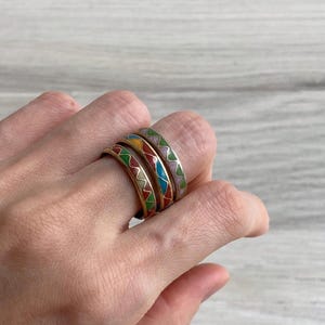 Puede incluir: Tres anillos de metal apilados con un patrón en zigzag relleno de esmalte de colores. Cada banda muestra un esquema de color distinto, incluyendo tonos de rojo, verde, azul, amarillo y morado, engastados en un marco metálico.