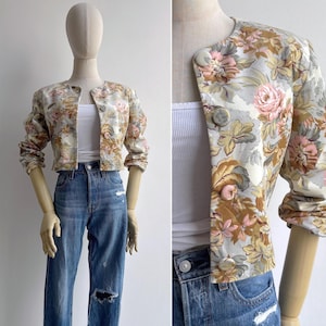 May include: A cropped jacket featuring a floral pattern in shades of pink, tan, and grey on a cream background. The jacket has a round neckline, long sleeves, and a front button closure with four large, round, matching fabric-covered buttons.