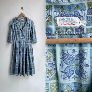May include: Vintage blue and green patterned dress with a collared neckline and three-quarter sleeves. The dress features a button-down front and a pleated skirt. The tag reads "TANNER of North Carolina by Dorothy Cox".