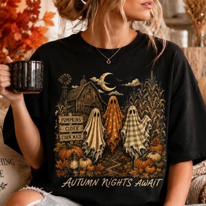 Op de afbeelding: Een zwart t-shirt met een afbeelding van drie geesten in een veld bij een schuur en een halve maan. Het ontwerp bevat de tekst PUMPKINS, CIDER, CORN MAZE en AUTUMN NIGHTS AWAIT, samen met pompoenen en een lantaarn.