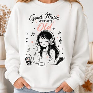 Op de afbeelding: Een wit sweatshirt met lange mouwen en een afbeelding van een persoon met lang haar die een koptelefoon draagt. De tekst luidt Good Music NEVER GETS Old. Het ontwerp bevat muzieknoten, kleine hartjes, een draagbare muziekspeler en een microfoon.