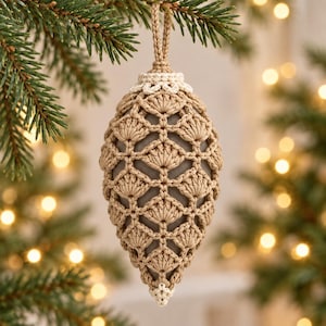 May include: A pinecone-shaped Christmas ornament featuring a dark gray core encased in a beige crocheted shell with a shell-stitch pattern. The ornament includes a matching beige crocheted hanging loop at the top and a small white crocheted tip.