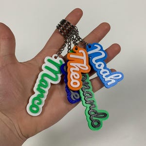 May include: A collection of five custom 3D-printed name keychains attached to a single metal ring. The keychains feature names including Marco, Theo, Noah, and Fernando in various colors such as green, orange, blue, and white with script lettering.
