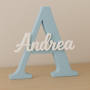 Könnte beinhalten: Ein hellblaues Holzbuchstaben "A" mit dem Namen "Andrea" in weißer Schrift. Der dekorative Buchstabe steht auf einer hellbraunen Oberfläche, mit einem neutralen Hintergrund.