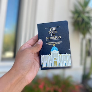 May include: A navy blue book titled "THE BOOK OF MORMON" with gold lettering and an illustration of a white building with a blue dome. The book is held in a hand.
