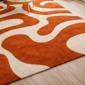 May include: An orange and cream-colored area rug with a wavy, abstract design. The rug is made of a soft, plush material and has a low pile. The rug is placed on a hardwood floor.