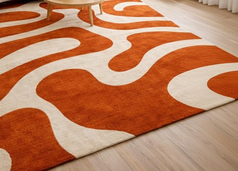 May include: An orange and cream-colored area rug with a wavy, abstract design. The rug is made of a soft, plush material and has a low pile. The rug is placed on a hardwood floor.