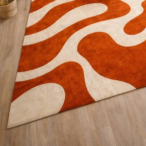 May include: An orange and cream-colored area rug with a wavy pattern. The rug is triangular in shape and has a soft, textured appearance. The design features broad, flowing cream-colored lines against a vibrant orange background. The rug is placed on a light wood floor.