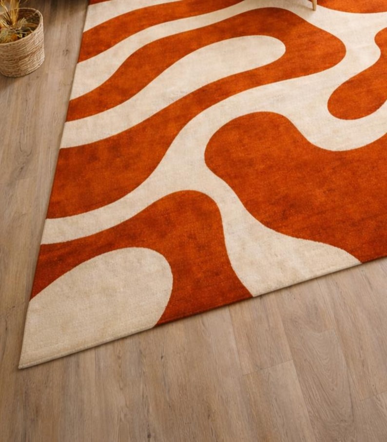 May include: An orange and cream-colored area rug with a wavy pattern. The rug is triangular in shape and has a soft, textured appearance. The design features broad, flowing cream-colored lines against a vibrant orange background. The rug is placed on a light wood floor.