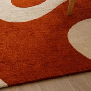 May include: A close-up of a rug with a bold, abstract design. The rug features a burnt orange background with a flowing, cream-colored pattern. The texture appears soft and plush. A wooden chair leg is visible.