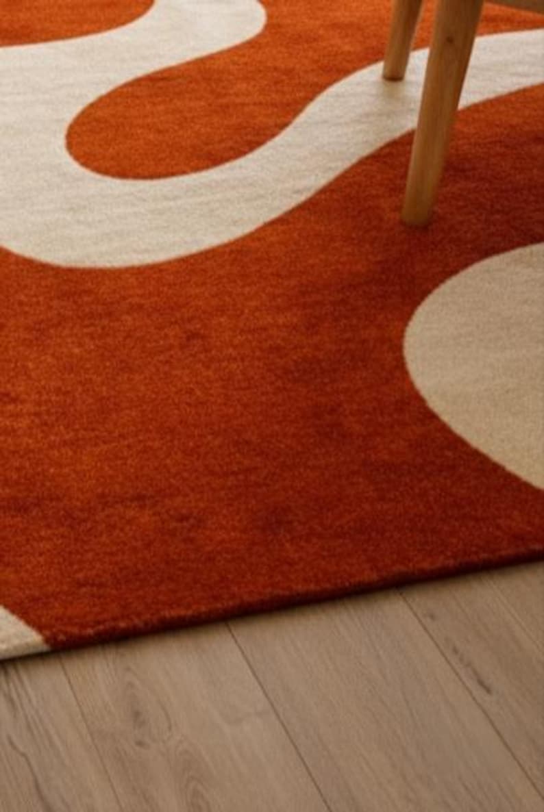 May include: A close-up of a rug with a bold, abstract design. The rug features a burnt orange background with a flowing, cream-colored pattern. The texture appears soft and plush. A wooden chair leg is visible.